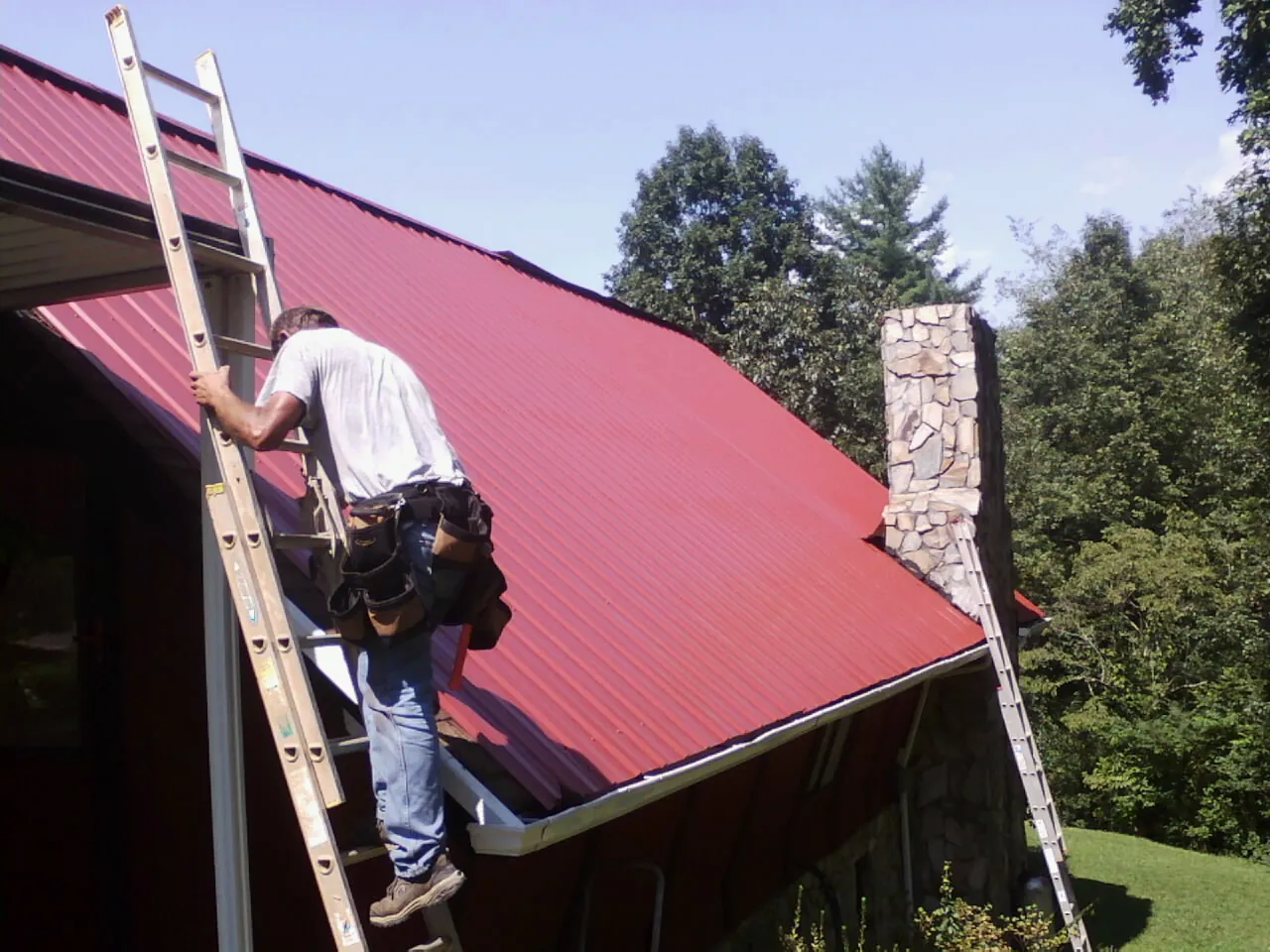 Roofer on a ladder beside a red metal roof and stone chimney — Soffit & Fascia Repair by Roofing Brothers in Valley Lee